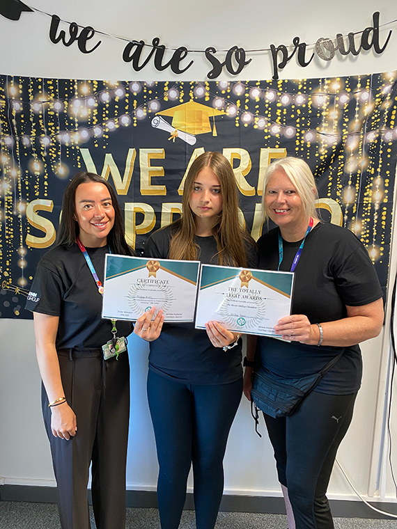 Three females stood together holding two certificates, two banners behind them saying 'we are so proud'