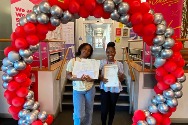 Two smiling females each holding up a piece of paper, stood at the foot of some stairs under a red and silver balloon arch