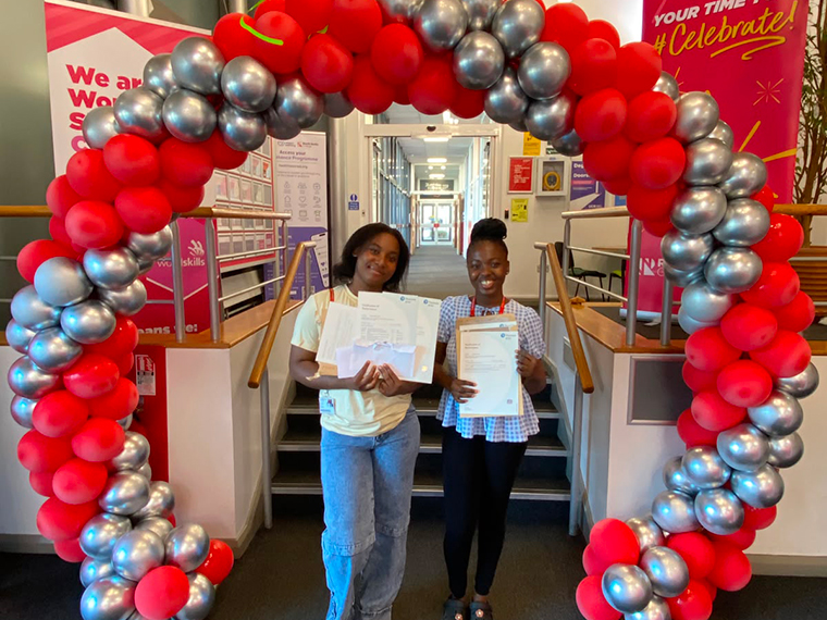 Two smiling females each holding up a piece of paper, stood at the foot of some stairs under a red and silver balloon arch