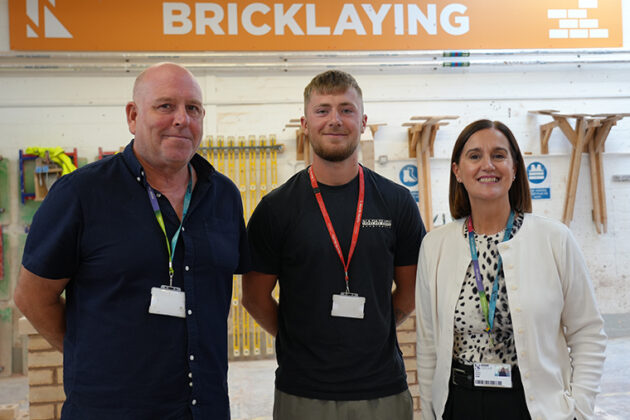 Two males and a female stood together, an orange and white sign above them that says 'Bricklaying'