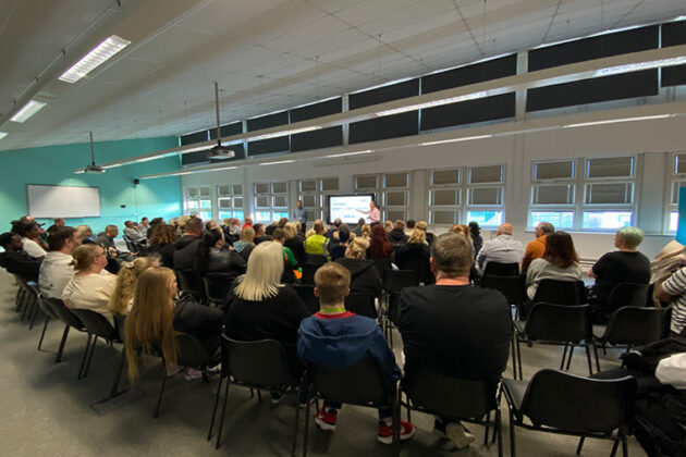 A room full of seated people looking towards a television screen and two people stood either side it presenting to the room