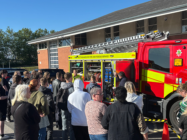 A fire engine with the side doors off showing the equipment that it carries, a crowd of people listening to someone speak about it