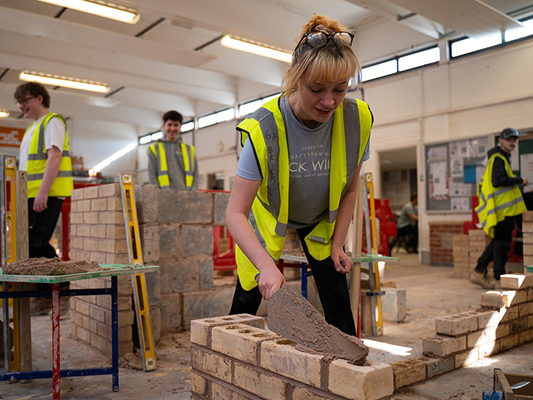 A female with blonde hair in a pony tail wearing a high jacket building a brick wall inside a workshop