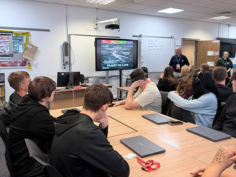 A group of people sat around tables in a room looking towards a screen at the front that says 'Float To Live'