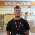 A male wearing a black tee shirt and a red lanyard stood with his hands behind his back, an orange and white sign reading 'Bricklaying' on the wall behind him