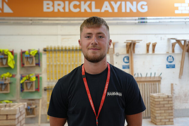 A male wearing a black tee shirt and a red lanyard stood with his hands behind his back, an orange and white sign reading 'Bricklaying' on the wall behind him