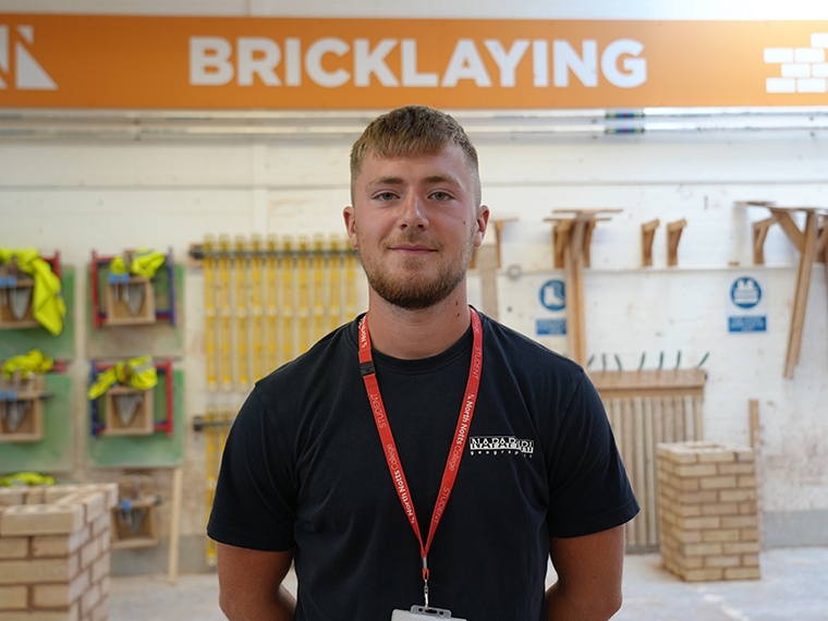 A male wearing a black tee shirt and a red lanyard stood with his hands behind his back, an orange and white sign reading 'Bricklaying' on the wall behind him