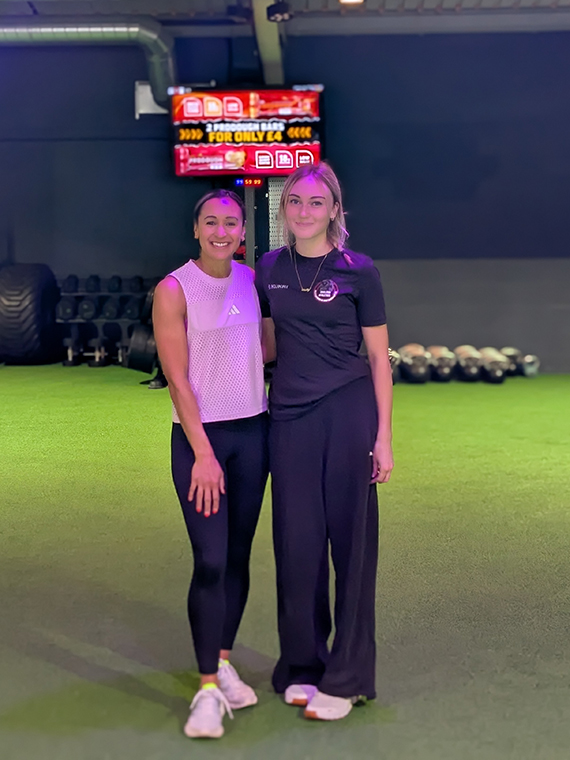 Two females, one blonde one brunette, stood side by side in a sports arena
