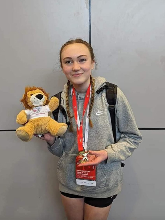 A smiling female wearing a light grey jumper, black shorts and a read lanyard, holding a teddy bear