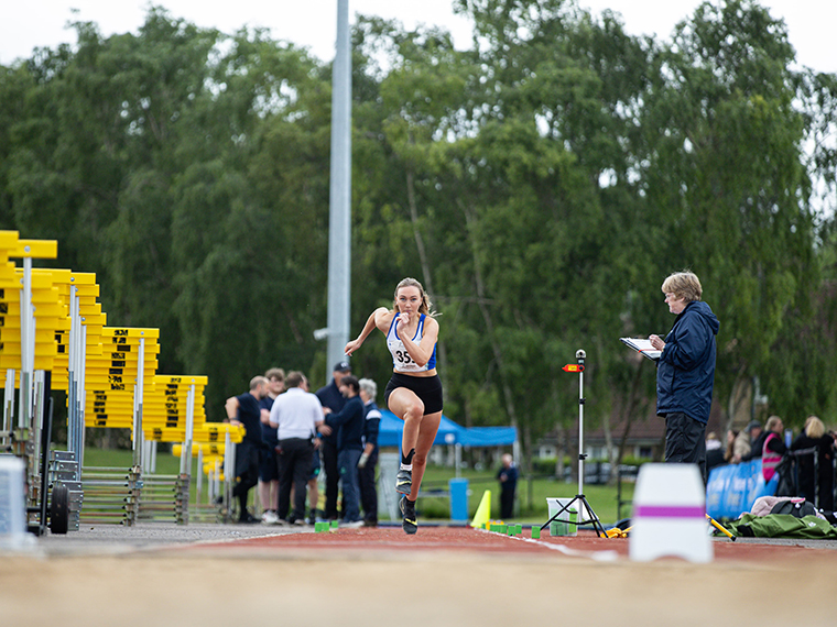 A female on a running track running towards the camera, someone stood to the side with a clipboard