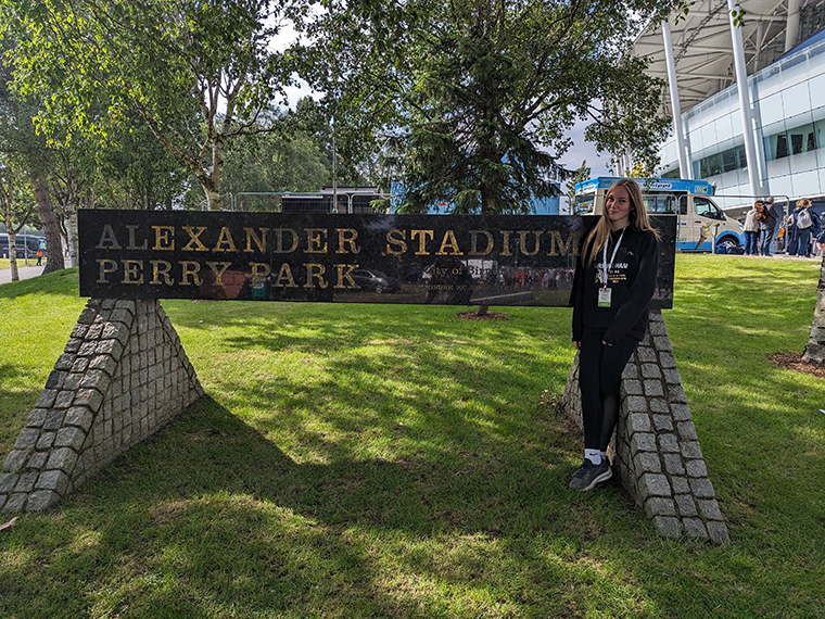 A blonde female wearing all black stood next to a large outdoor sign that reads 'Alexander Stadium Perry Park'