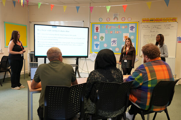 Three females stood next to a large white screen presenting to three seated people