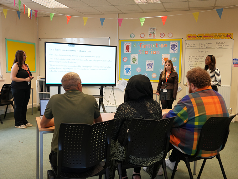 Three females stood next to a large white screen presenting to three seated people