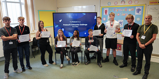 A group of people stood in a line in front of a large screen, three people seated in the middle, most are holding up a certificate